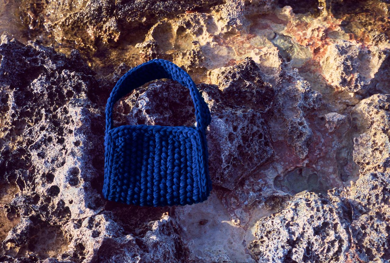 Bolso de ganchillo azul marino apoyado sobre roca porosa de la costa, con luz de atardecer
