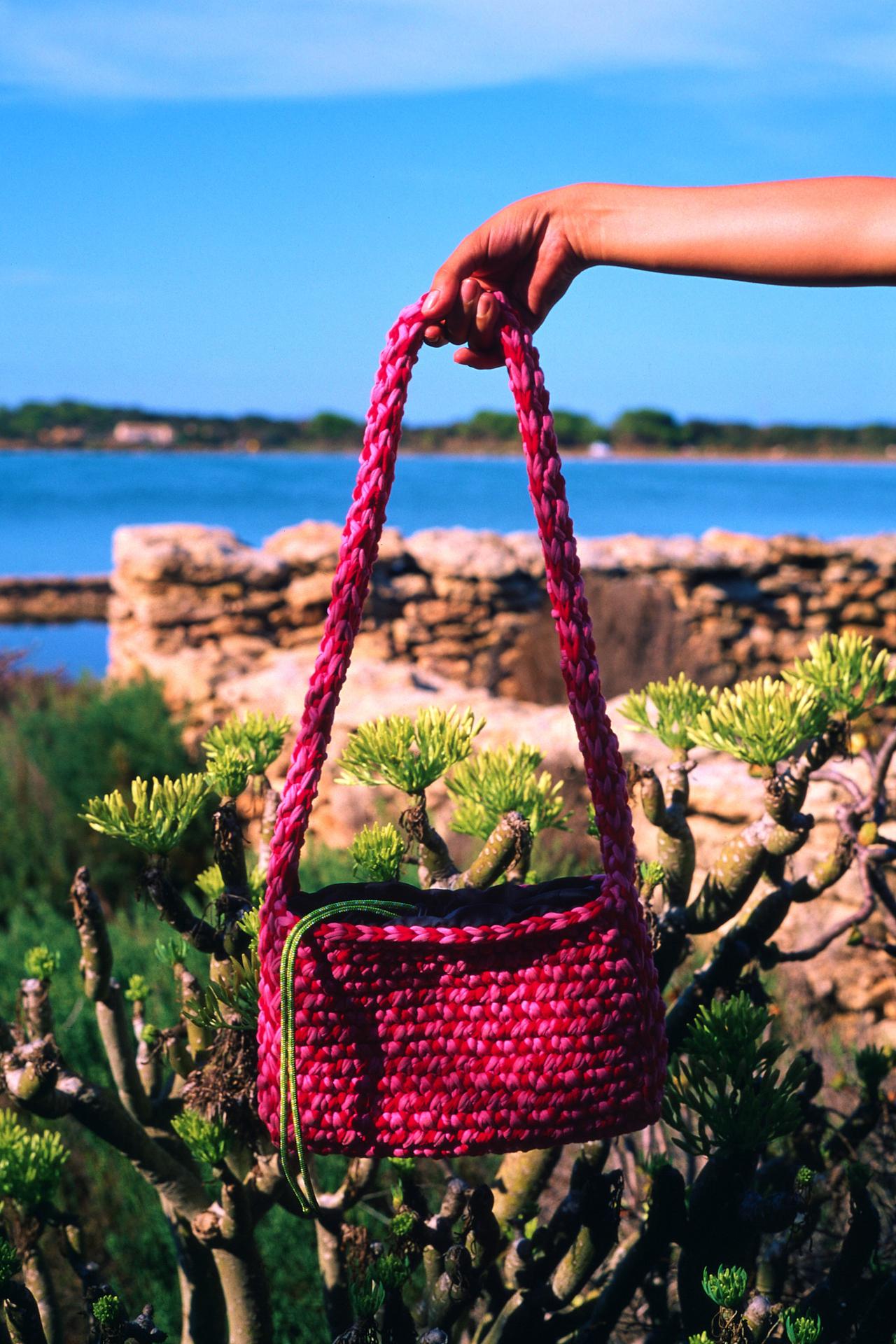 Mano sosteniendo un bolso de ganchillo fucsia, con el mar al fondo