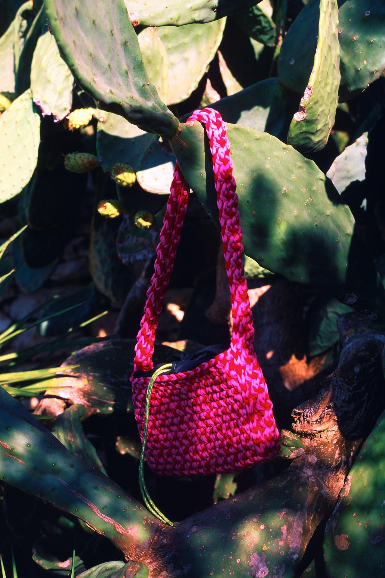 Bolso de ganchillo fucsia colgado entre las palas de una chumbera, a media sombra