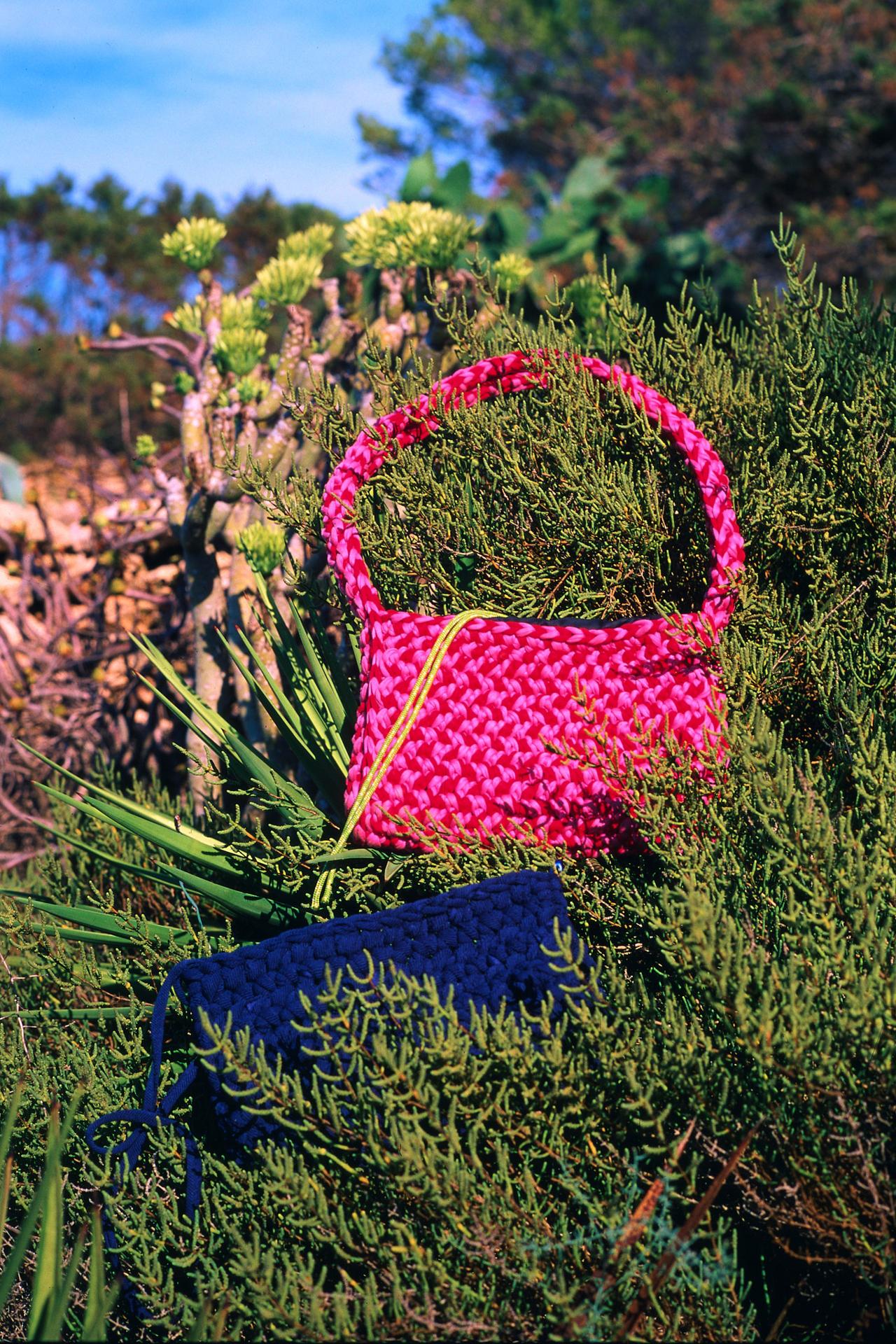 Bolso de ganchillo fucsia colgado de un arbusto, con un clutch azul marino debajo