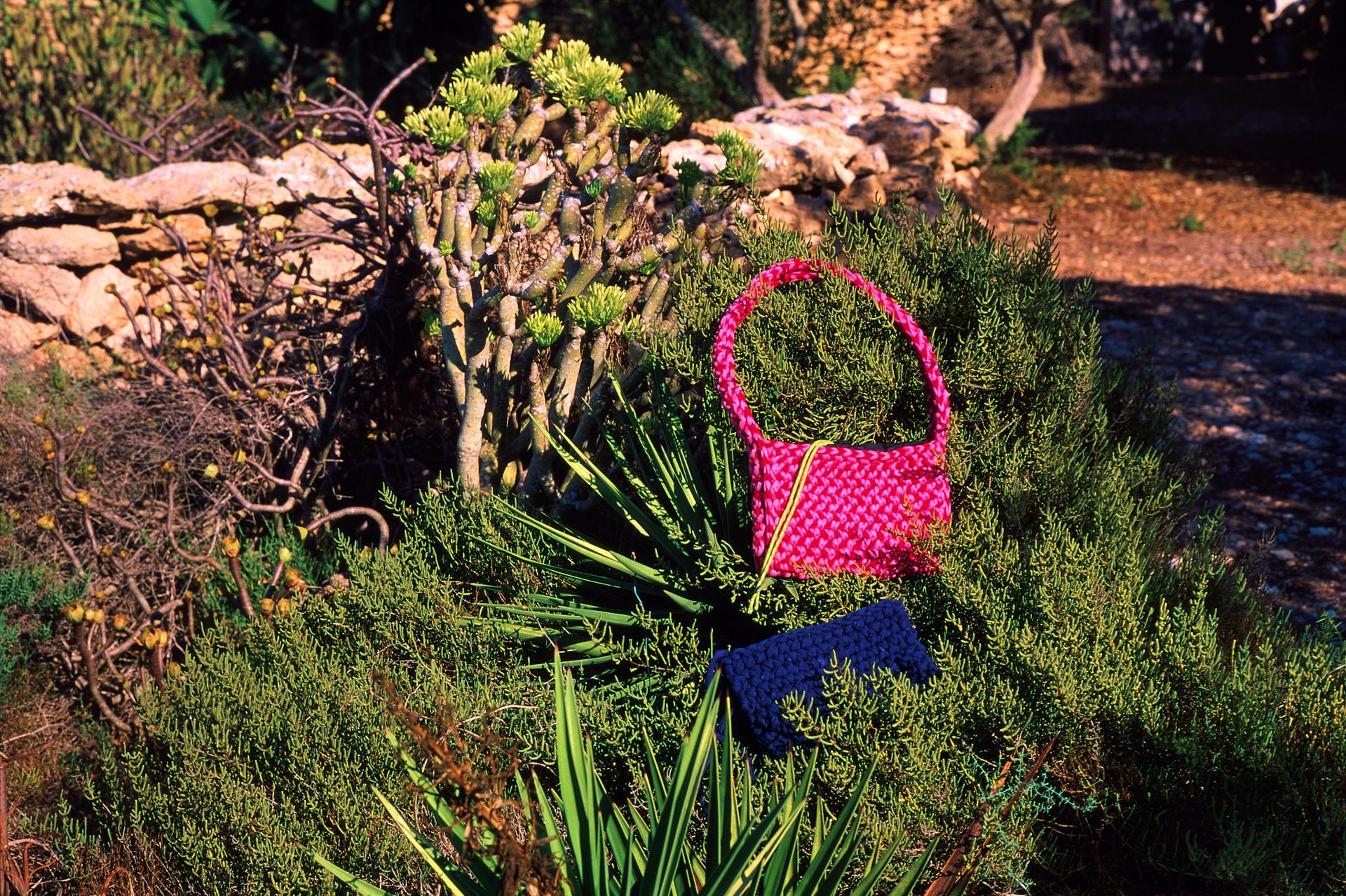Bolso de ganchillo fucsia y un clutch azul marino sobre matorral mediterráneo, junto a un muro de piedra seca