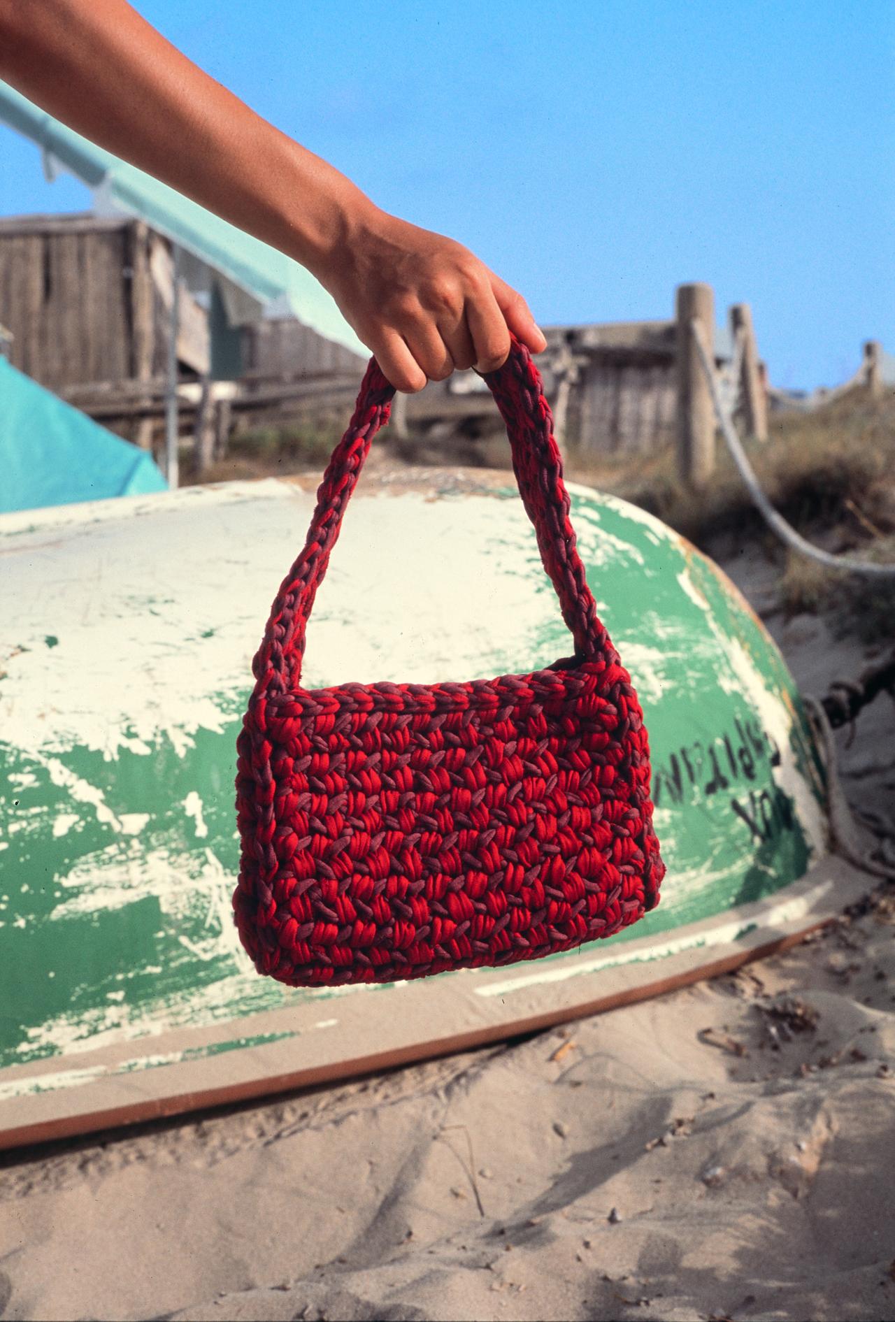 Mano sosteniendo un bolso de ganchillo rojo delante de una barca varada en la arena