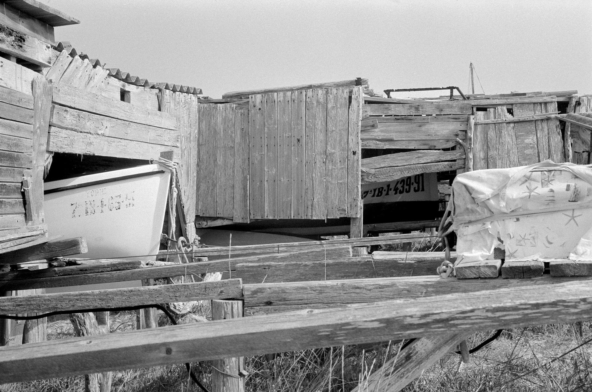 Casetas de madera y barcas guardadas bajo tablones junto a la orilla, en blanco y negro