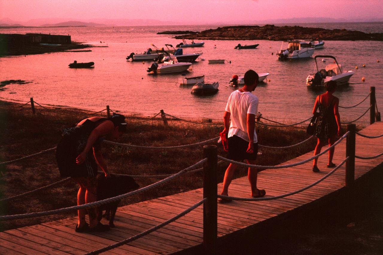 Gente paseando por una pasarela de madera junto a barcas fondeadas al atardecer