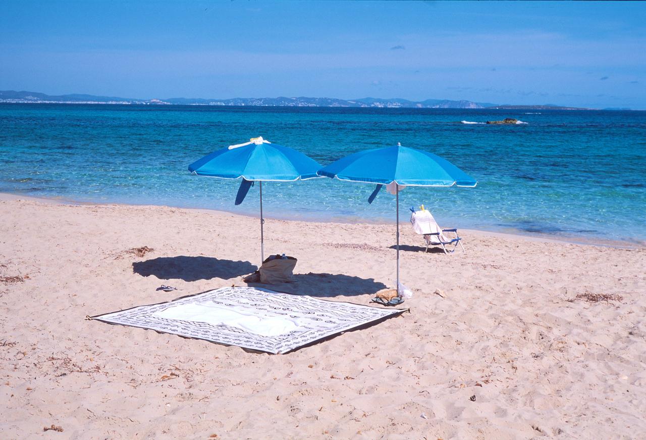 Dos sombrillas azules, una toalla y una silla en una playa de agua turquesa