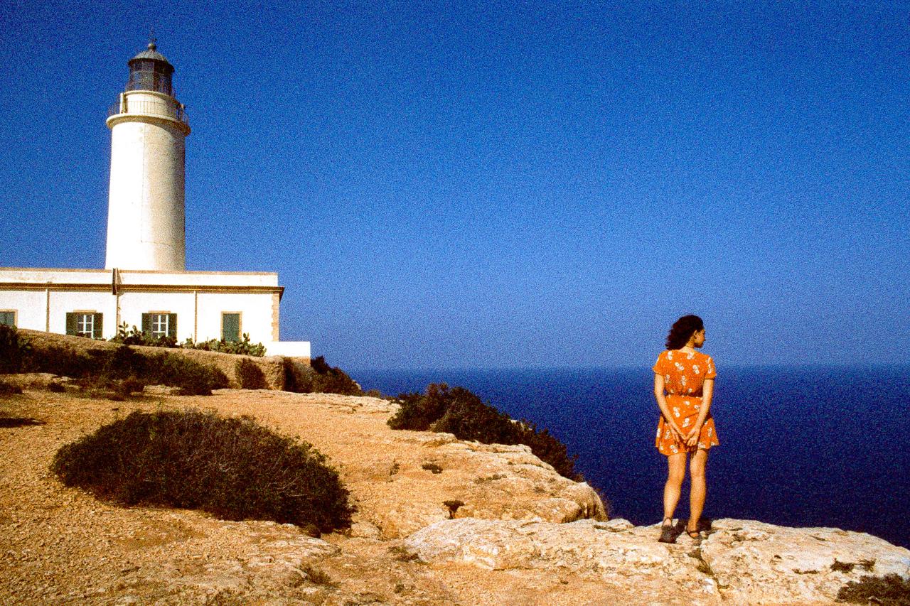 Mujer con vestido naranja de espaldas al borde de un acantilado, junto a un faro blanco