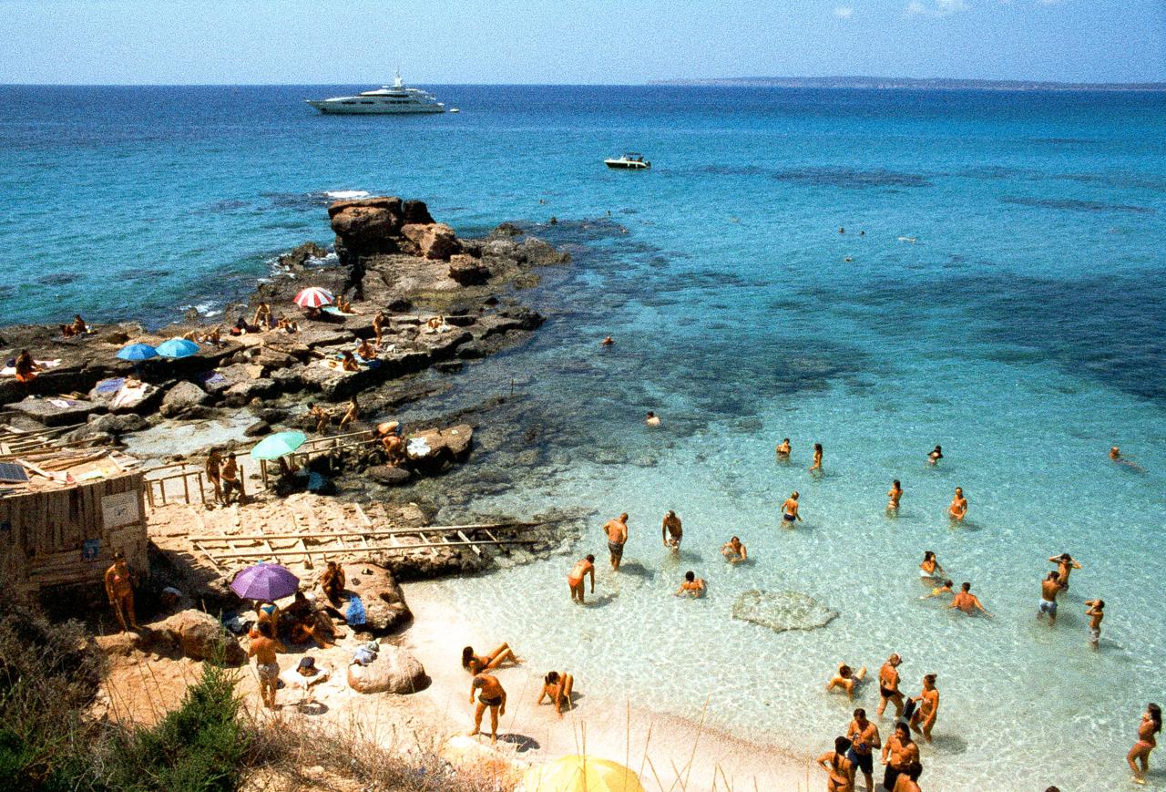 Bañistas en una cala de agua turquesa entre rocas, con un yate fondeado al fondo