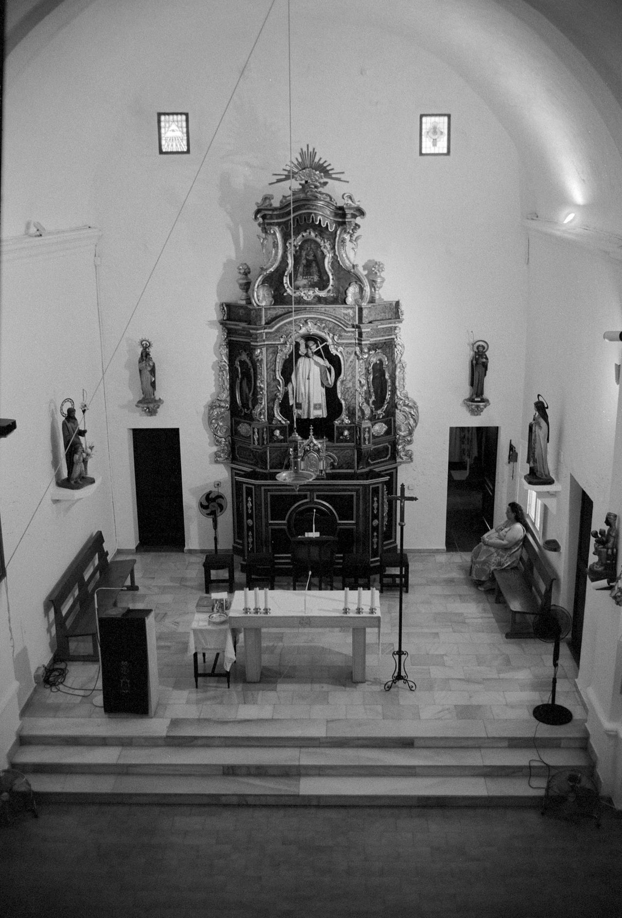 Interior de una iglesia encalada con un retablo barroco, visto desde el coro, en blanco y negro