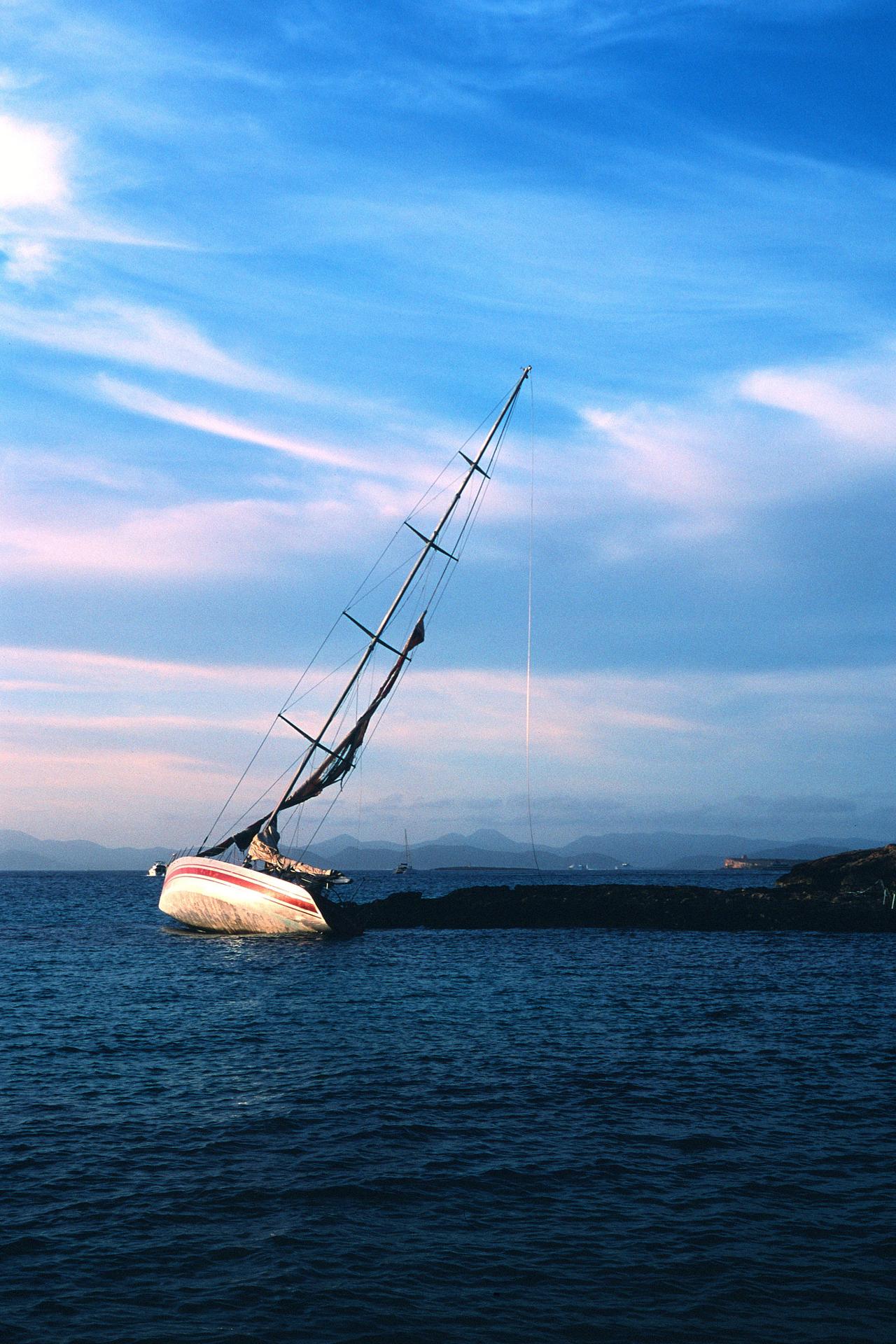 Velero escorado varado junto a unas rocas, bajo un cielo azul con nubes rosadas