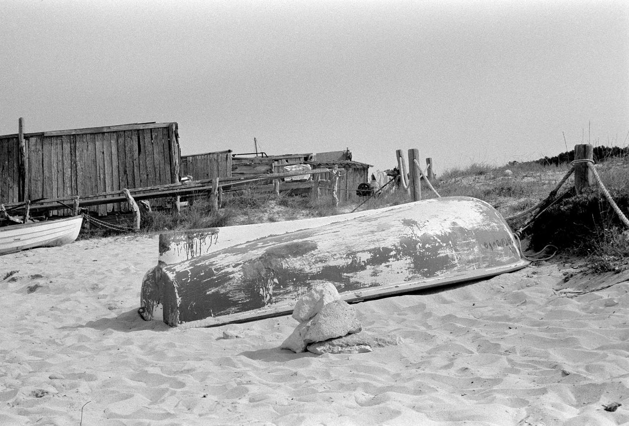 Barca de madera varada boca abajo en la arena junto a casetas de pescadores, en blanco y negro