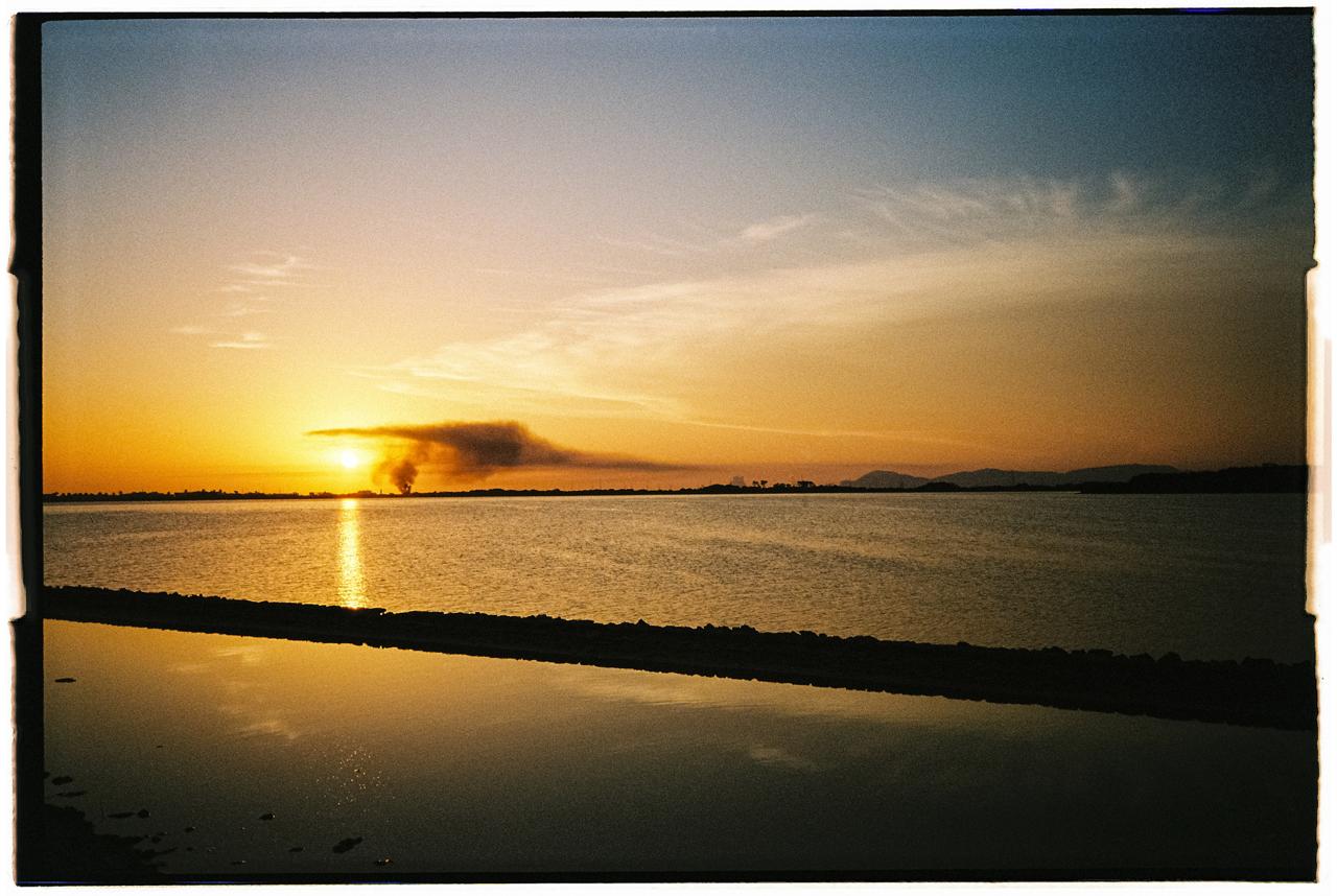 Puesta de sol sobre una laguna, con una columna de humo en el horizonte