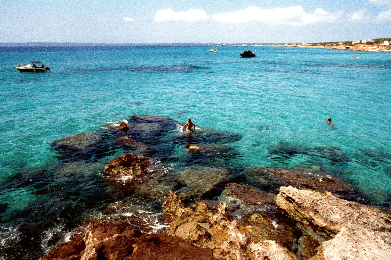 Rocas y agua turquesa transparente, con bañistas y barcas a lo lejos