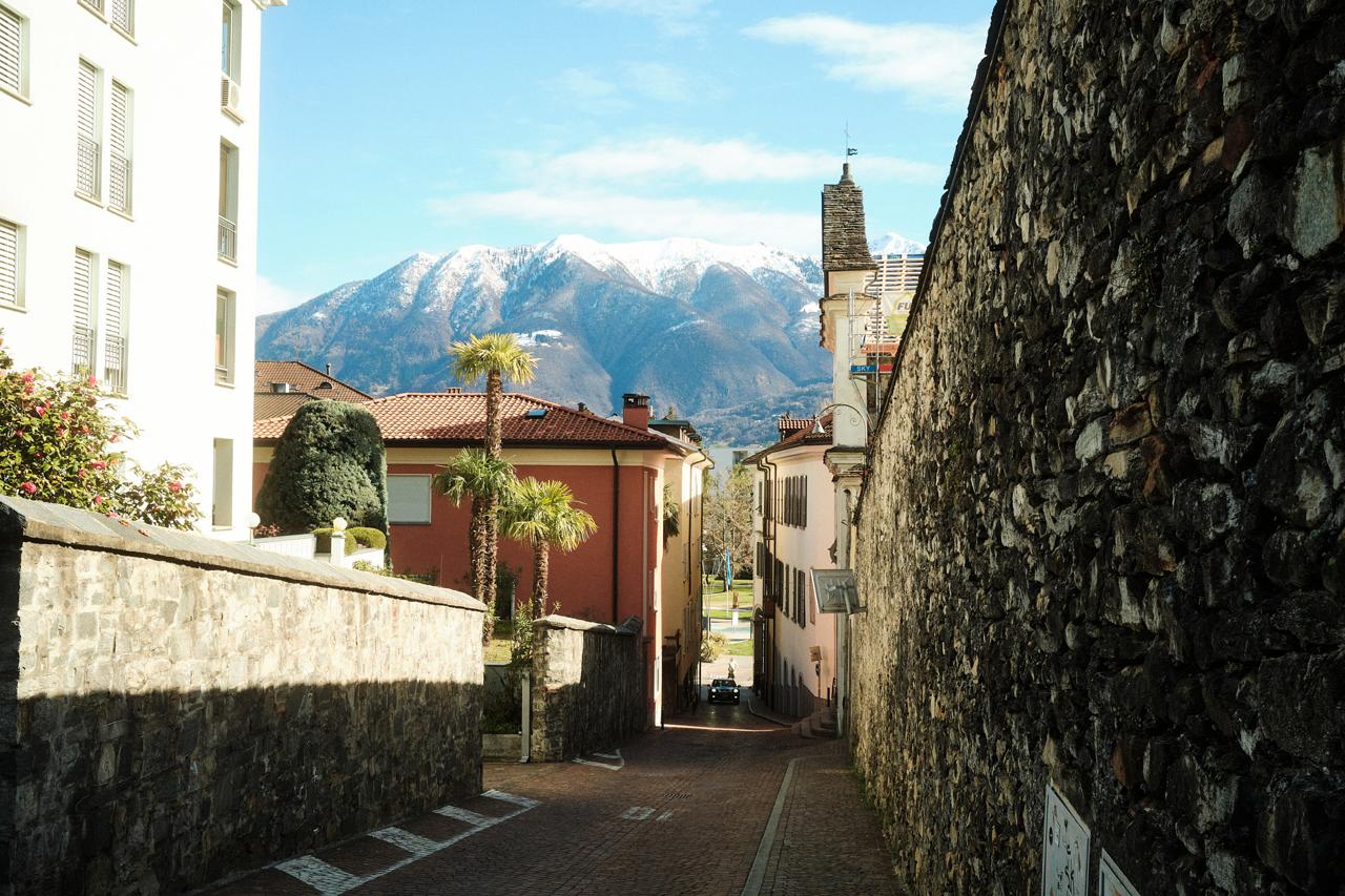 Calle empedrada entre muros de piedra, con palmeras y montañas nevadas al fondo