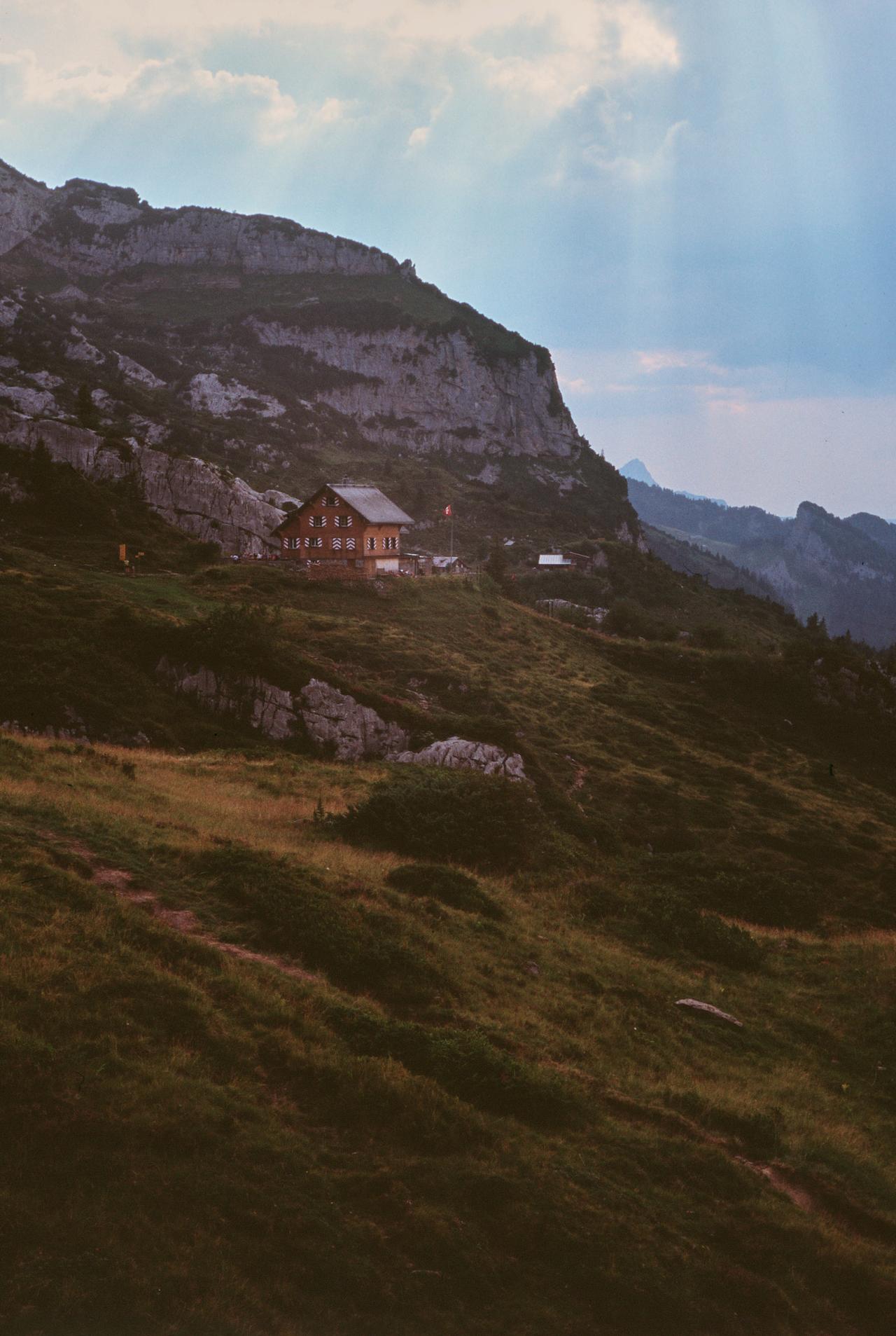 Refugio de montaña en una ladera bajo un acantilado, con rayos de sol entre las nubes
