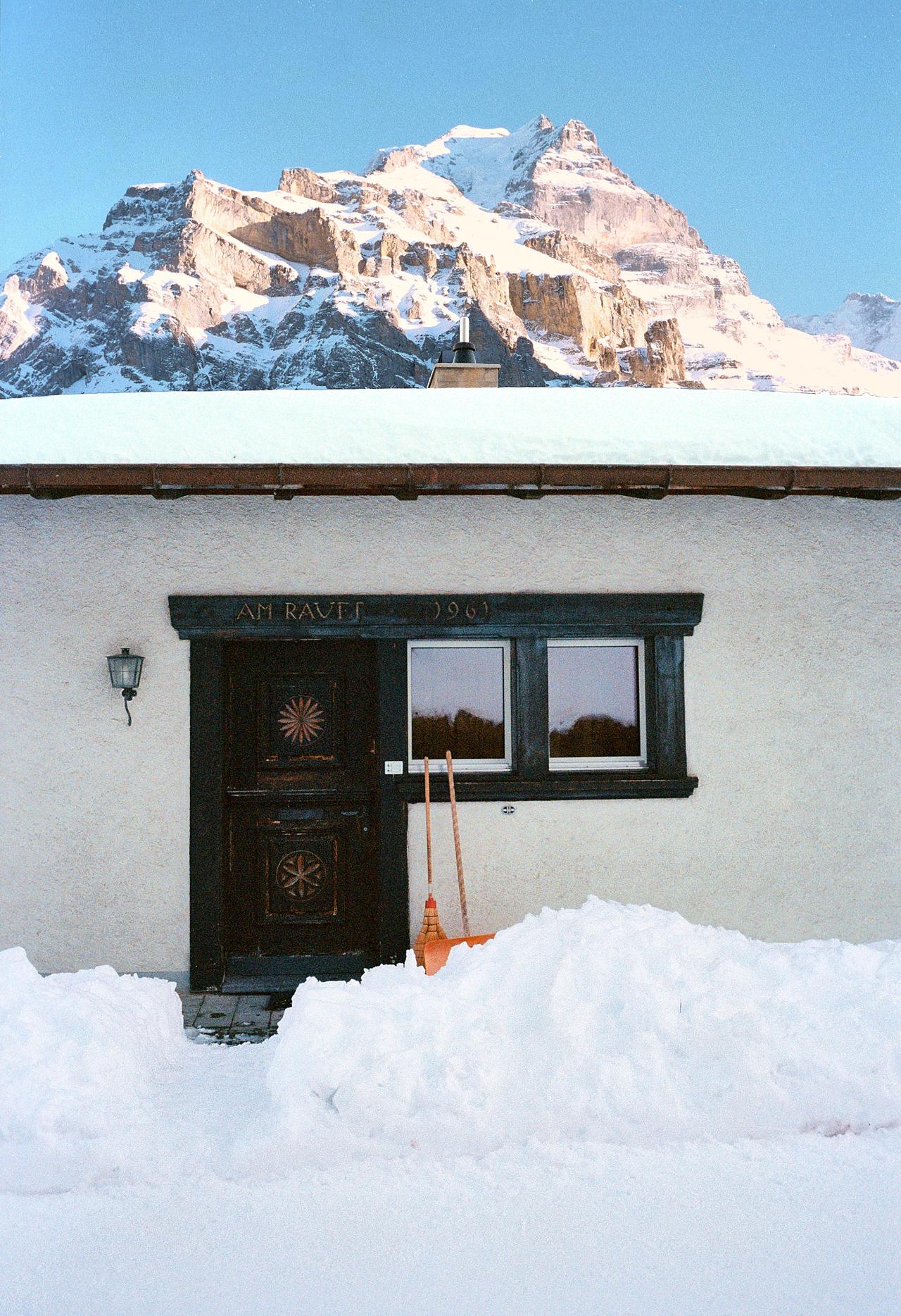 Casa encalada con la entrada despejada entre la nieve, bajo una montaña nevada
