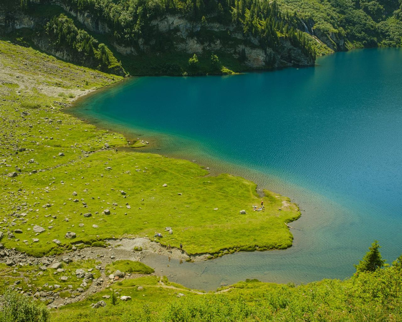 Lago alpino de agua turquesa bordeado por un prado verde con rocas, visto desde lo alto