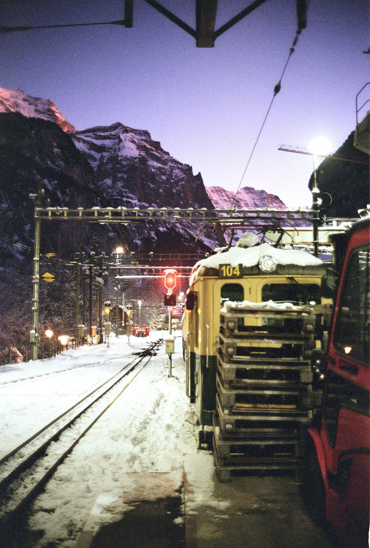 Estación de tren nevada de noche, con un vagón amarillo y montañas al fondo