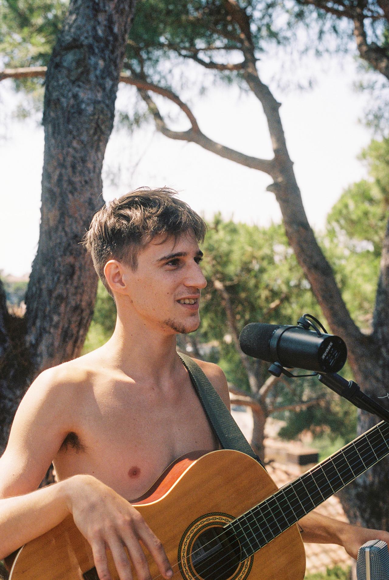 Joven sin camiseta con una guitarra acústica ante un micrófono, bajo unos pinos