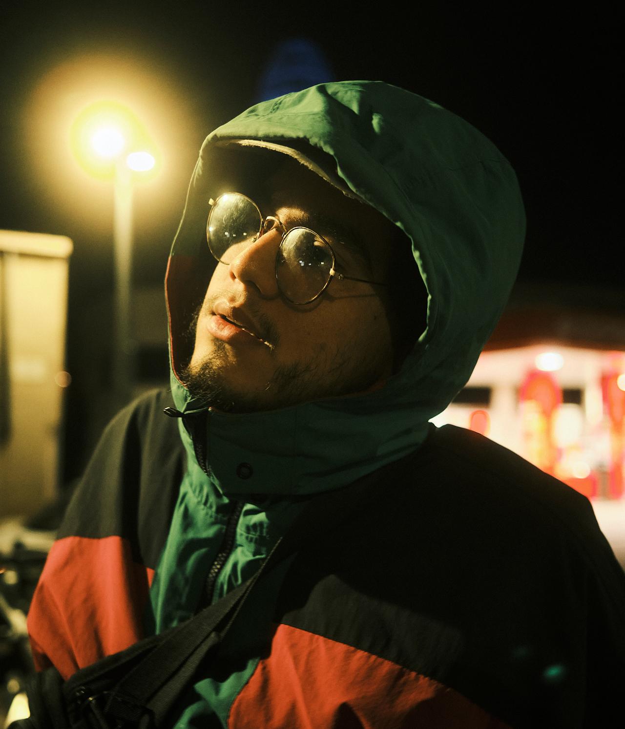 Joven con gafas redondas y capucha verde bajo una farola, de noche