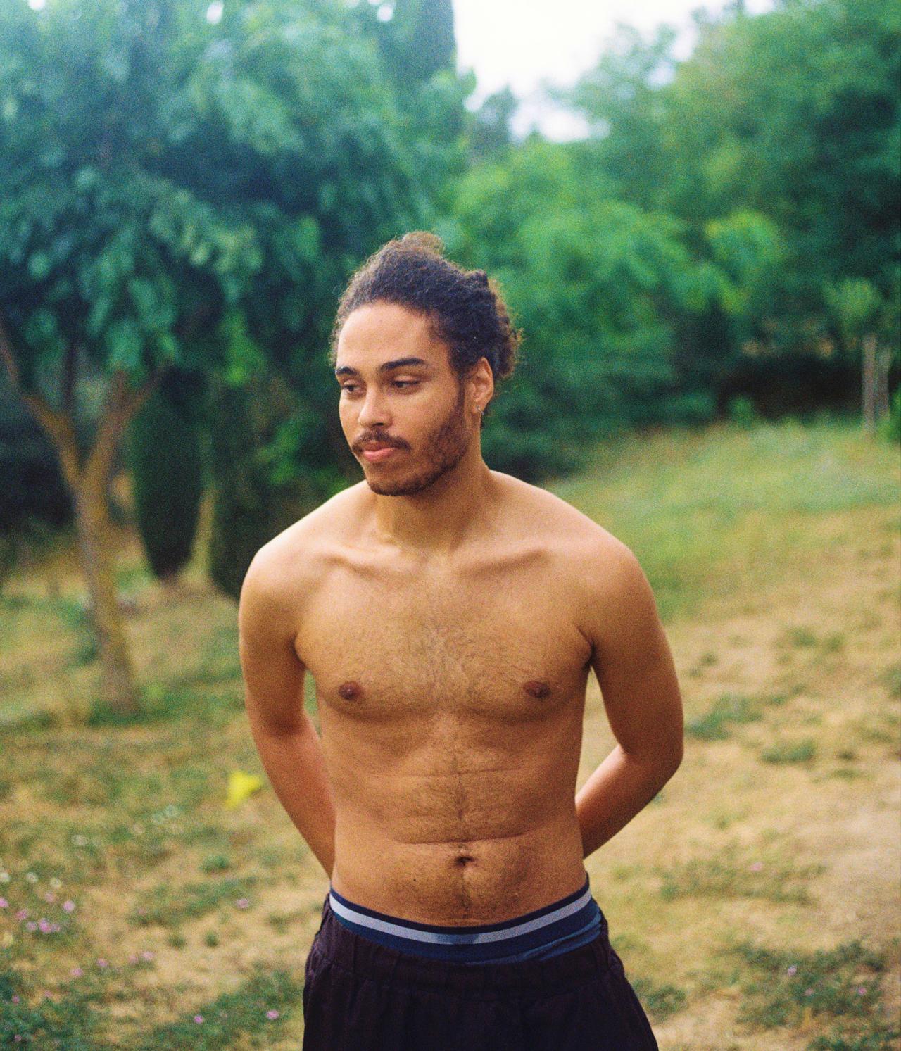 Joven sin camiseta con el pelo recogido en un moño, en un campo con árboles