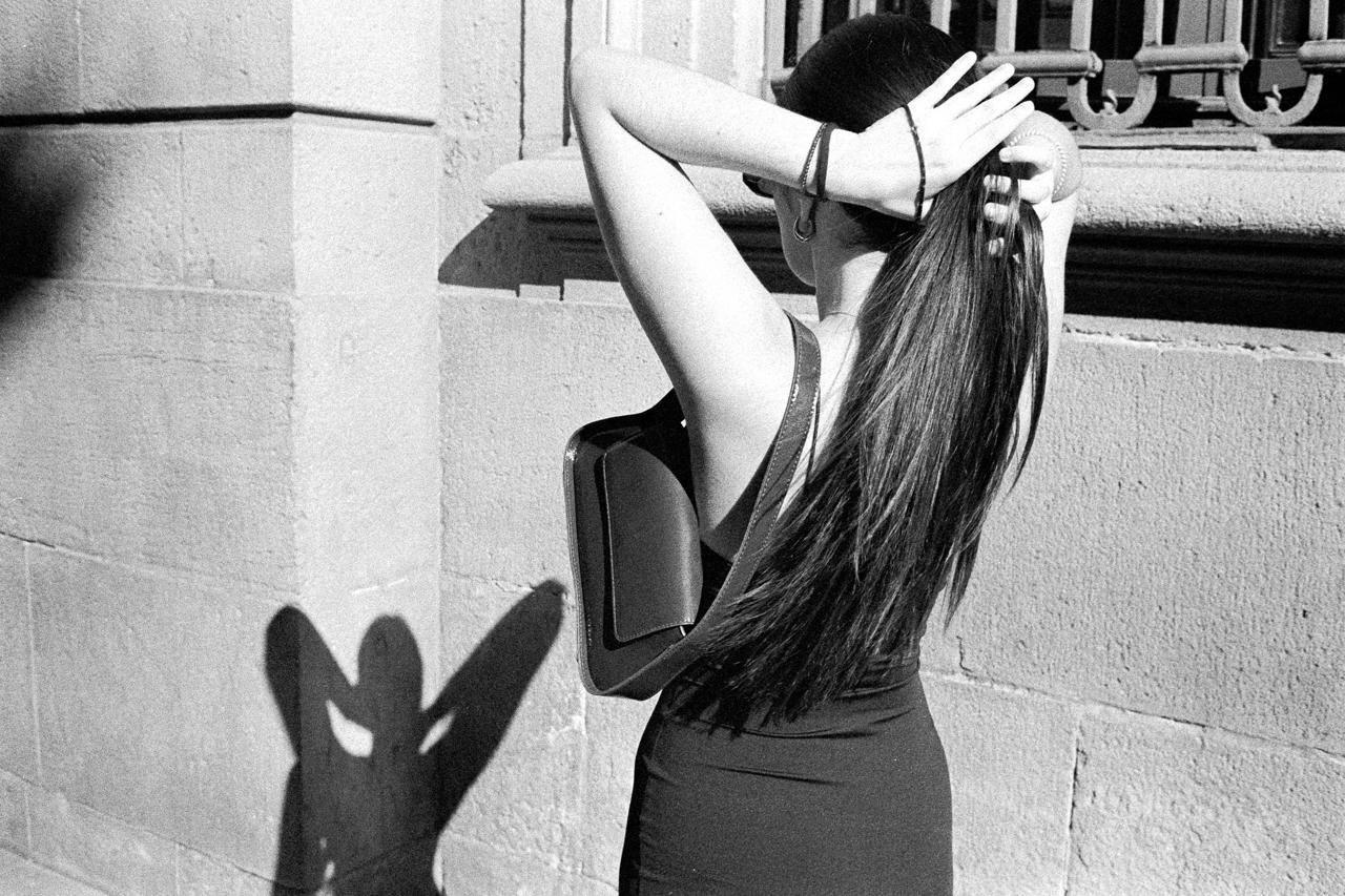 Mujer de espaldas recogiéndose el pelo junto a una fachada de piedra, en blanco y negro
