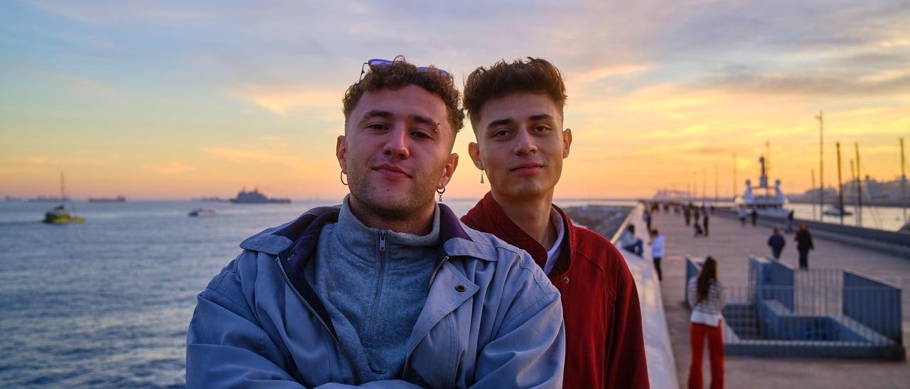 Dos jóvenes en un paseo marítimo al atardecer, con barcos y paseantes al fondo