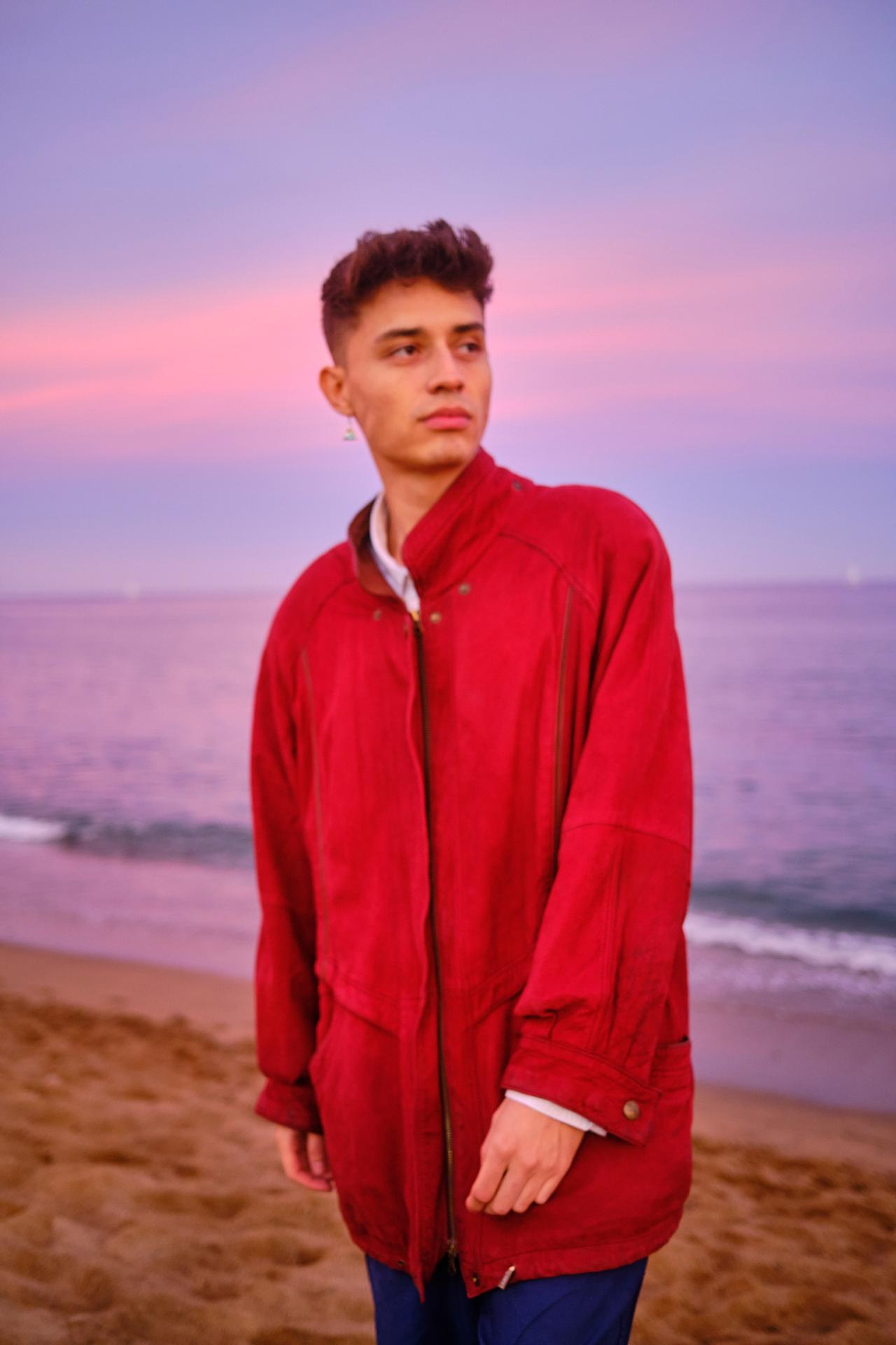 Joven con chaqueta roja en la orilla de una playa bajo un cielo rosado