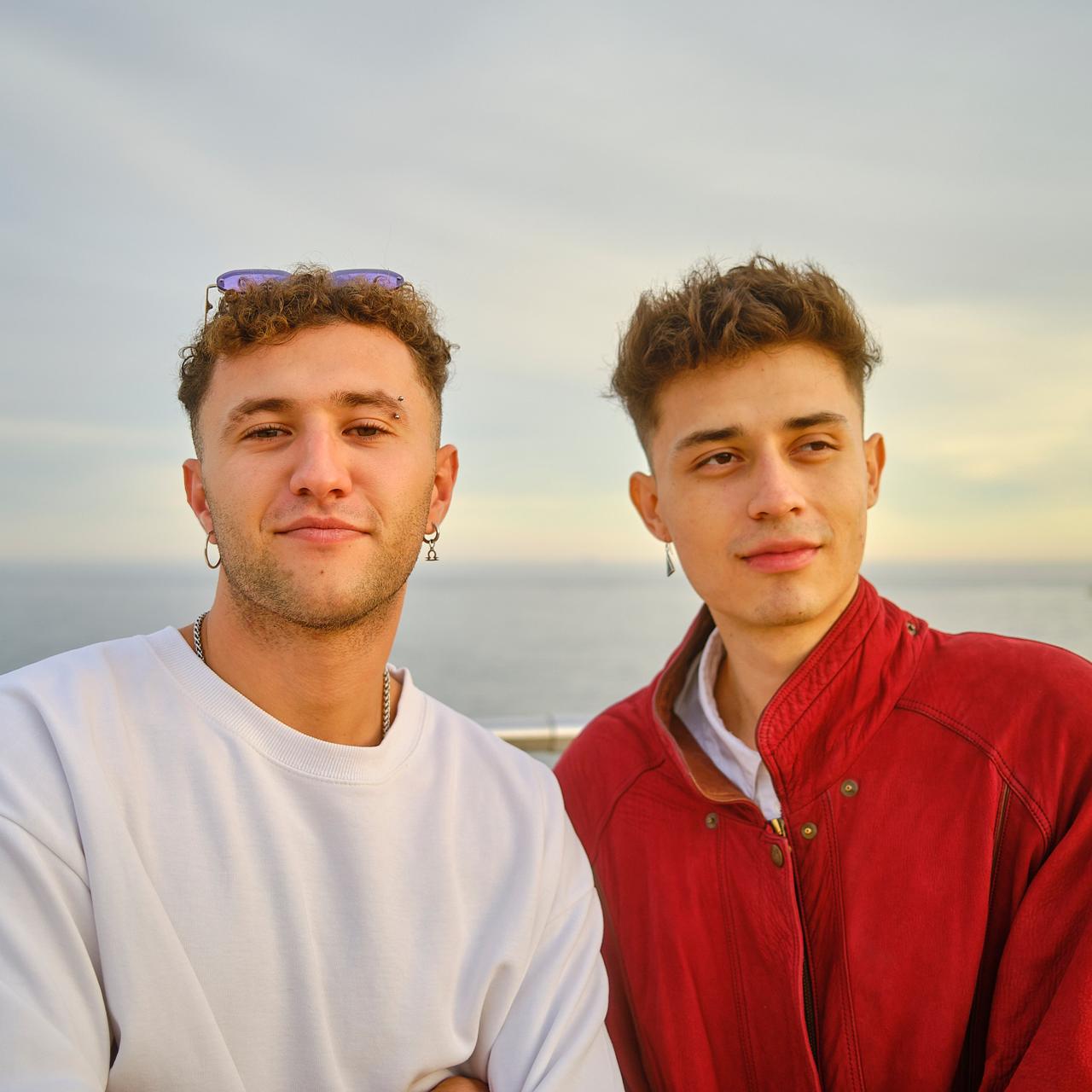 Dos jóvenes posando junto al mar al atardecer, uno con cazadora roja