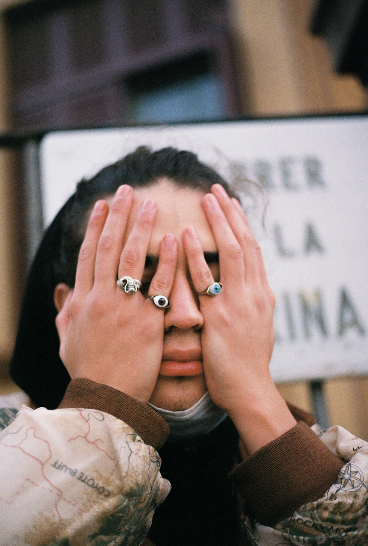 Persona tapándose los ojos con las manos llenas de anillos, delante de un cartel de calle