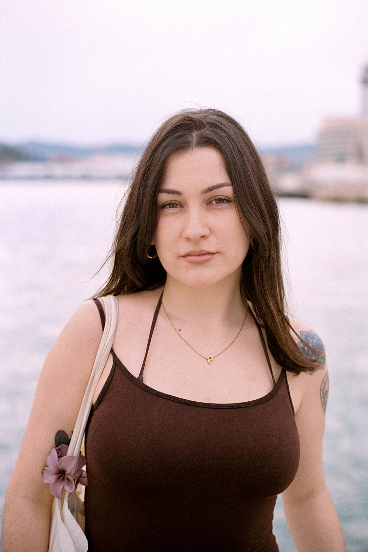 Mujer con camiseta marrón de tirantes junto al mar, con un puerto al fondo