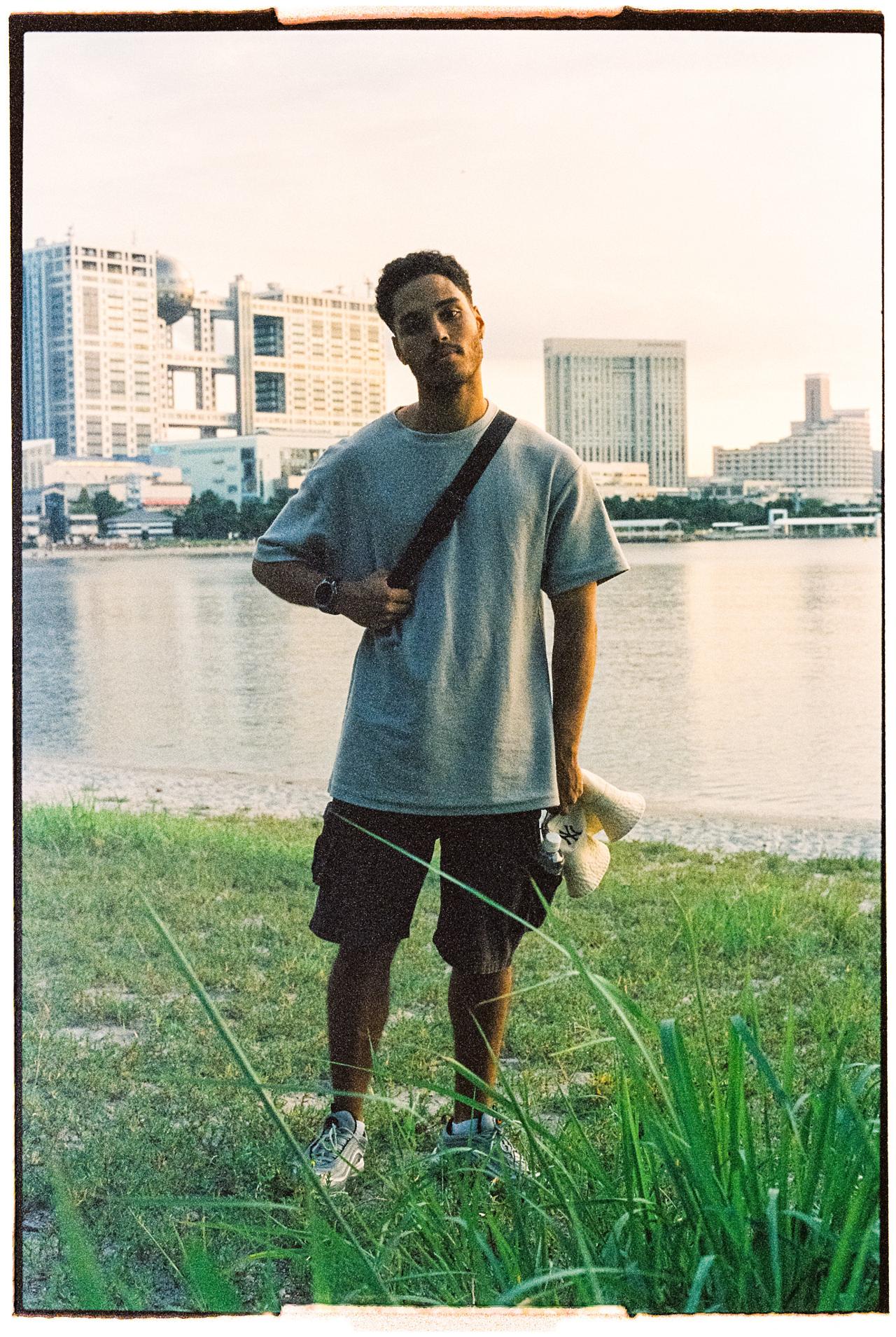 Joven con camiseta gris y bandolera junto al agua, con edificios de una ciudad al fondo