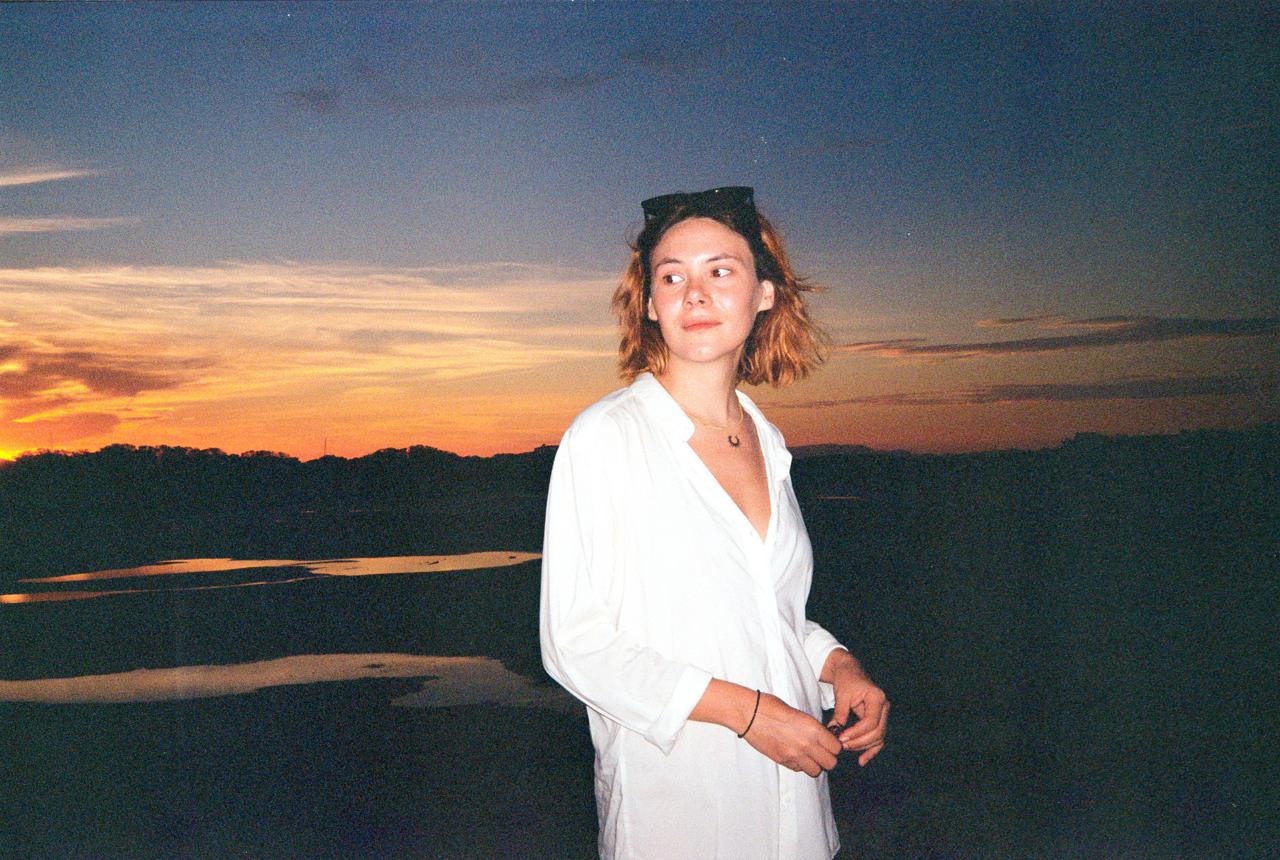 Mujer con camisa blanca y gafas de sol en la cabeza al anochecer, junto a una laguna