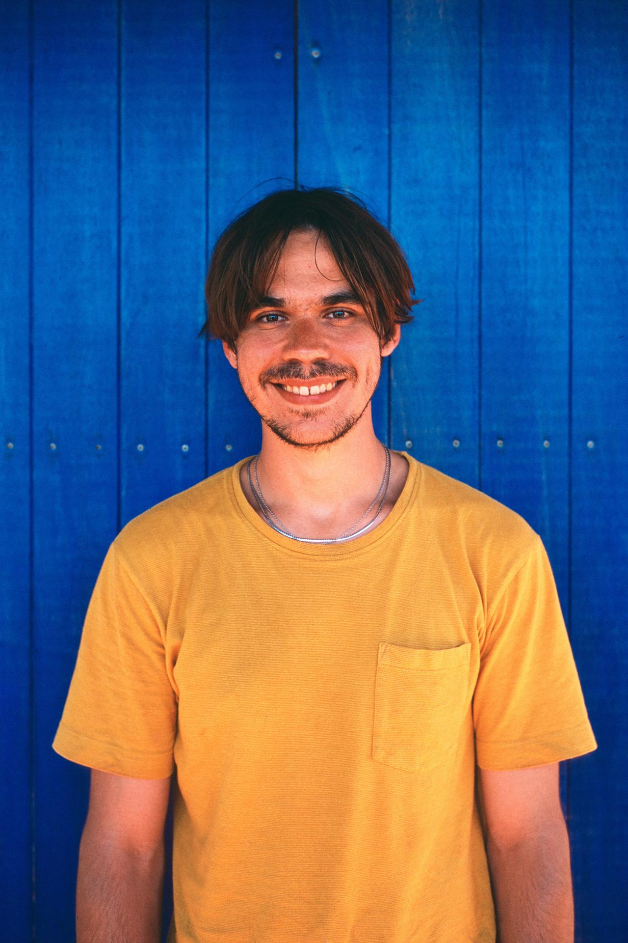 Hombre joven sonriente con camiseta amarilla frente a una puerta de madera azul