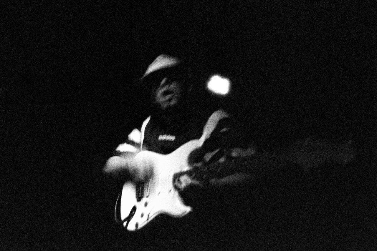 Guitarrista con gorra tocando una guitarra eléctrica blanca en la penumbra, en blanco y negro