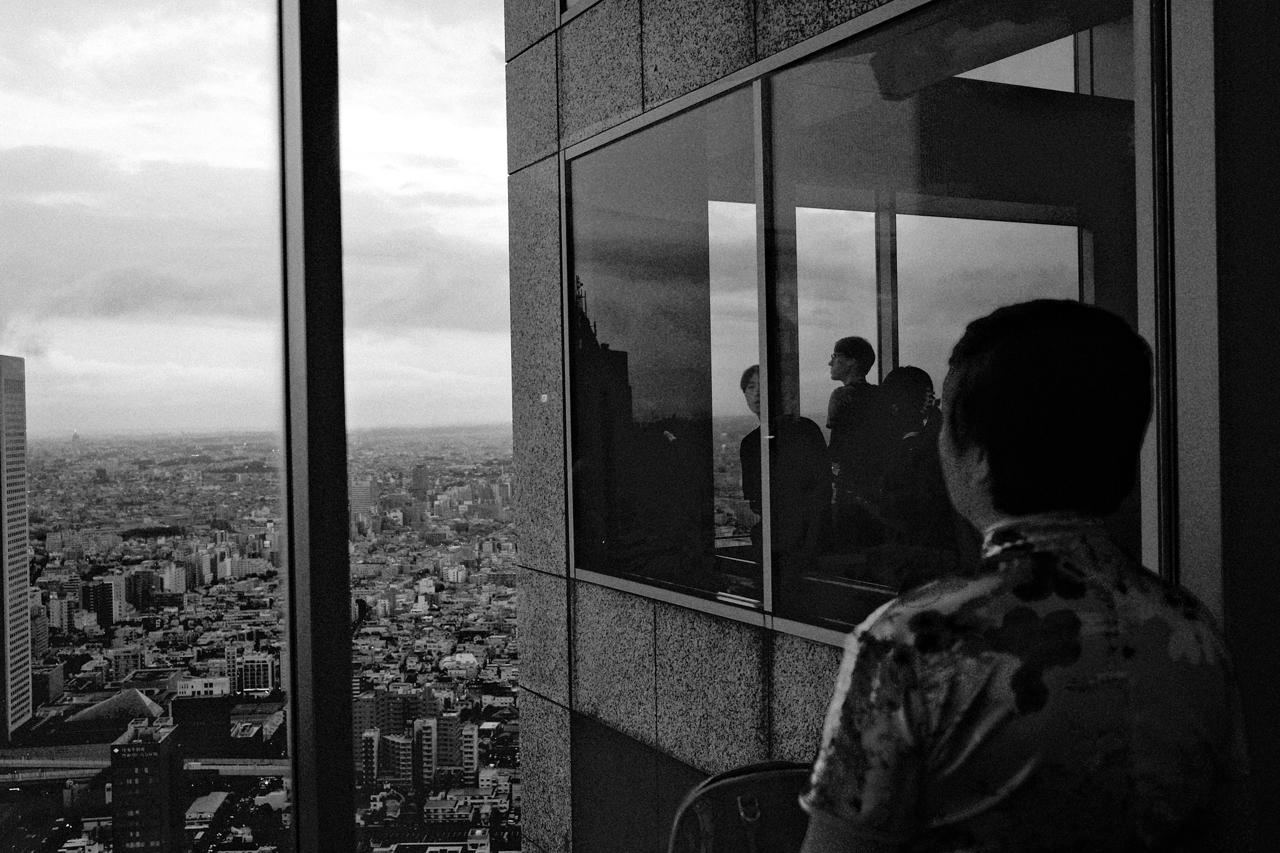 Hombre de espaldas mirando la ciudad desde un mirador, con otros visitantes reflejados en el cristal